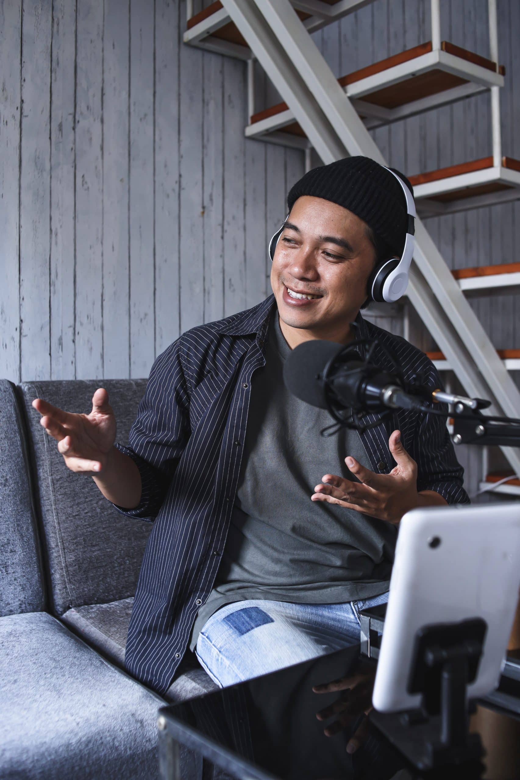 An Asian man is hosting a podcast. He is wearing a black beanie cap, black striped shirt, and white headphones.