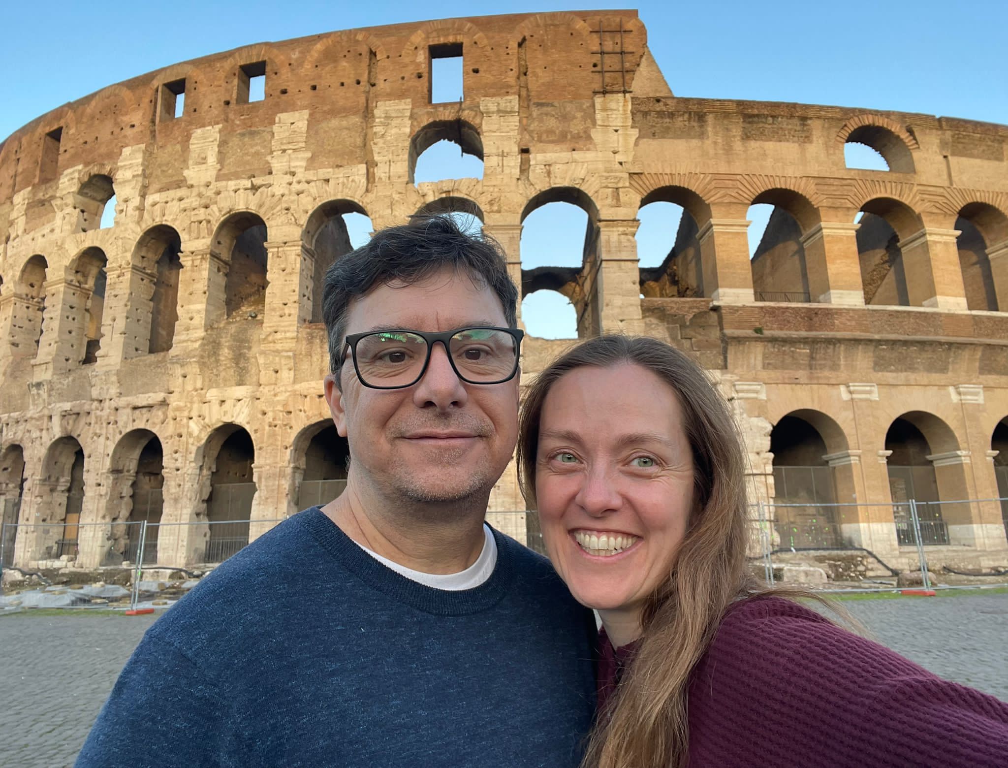 A man and woman in front of the Roman Colosseum. He is wearing a blue shirt and glasses. The woman has light brown hair and a maroon sweater